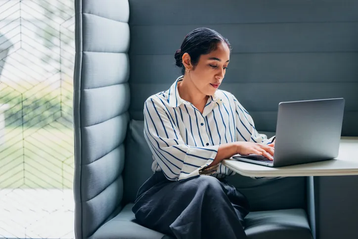 A professional woman sitting in a modern grey acoustic booth, typing on a laptop computer to illustrate digital work and online payment processing.