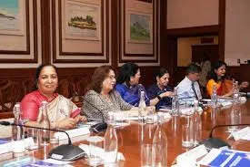 A group of women seated at a long wooden conference table with microphones and water bottles during a formal advisory meeting.