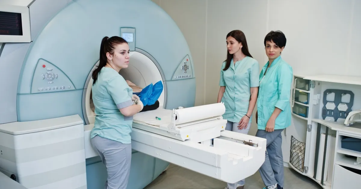 Three medical professionals in teal scrubs attending to a patient lying on the sliding bed of a large, modern medical scanner in a clinical hospital room.