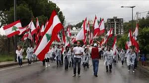 People wearing rain ponchos march down a wet street carrying large Lebanese flags during a national procession.