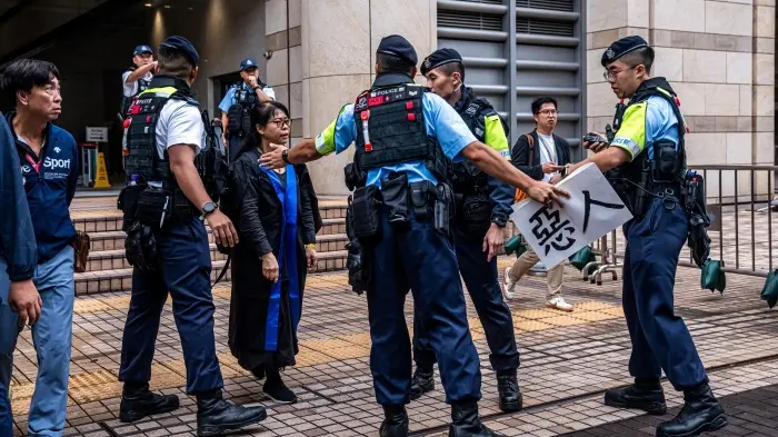 Uniformed Hong Kong police officers surround pro-democracy activist Alexandra Wong, known as Grandma Wong, outside the West Kowloon Magistrates' Courts as an officer confiscates a paper sign with the Chinese characters for 'Villain'.