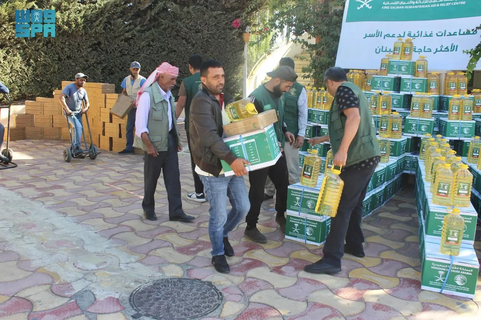 Men carrying green and white KSrelief food aid boxes and yellow cooking oil jugs in Tripoli, Lebanon, next to stacks of supplies and a banner.