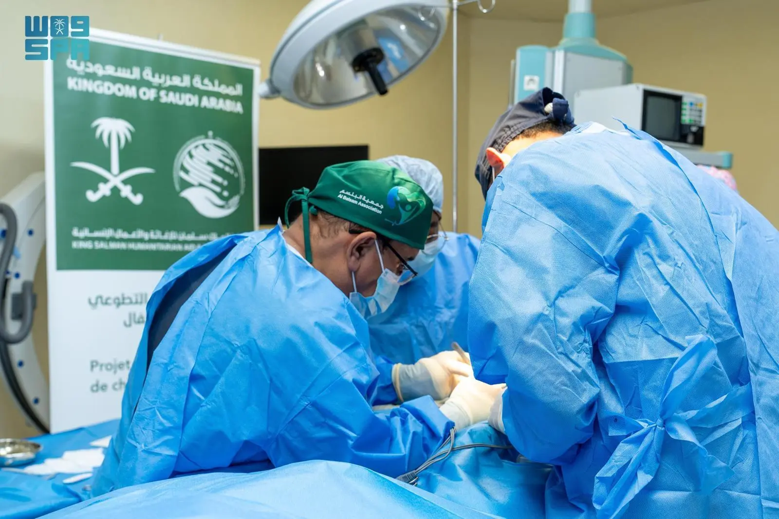 A team of surgeons from KSrelief and Al Balsam Association performing a medical procedure in an operating room in Moroni, Comoros, with a Kingdom of Saudi Arabia banner in the background.