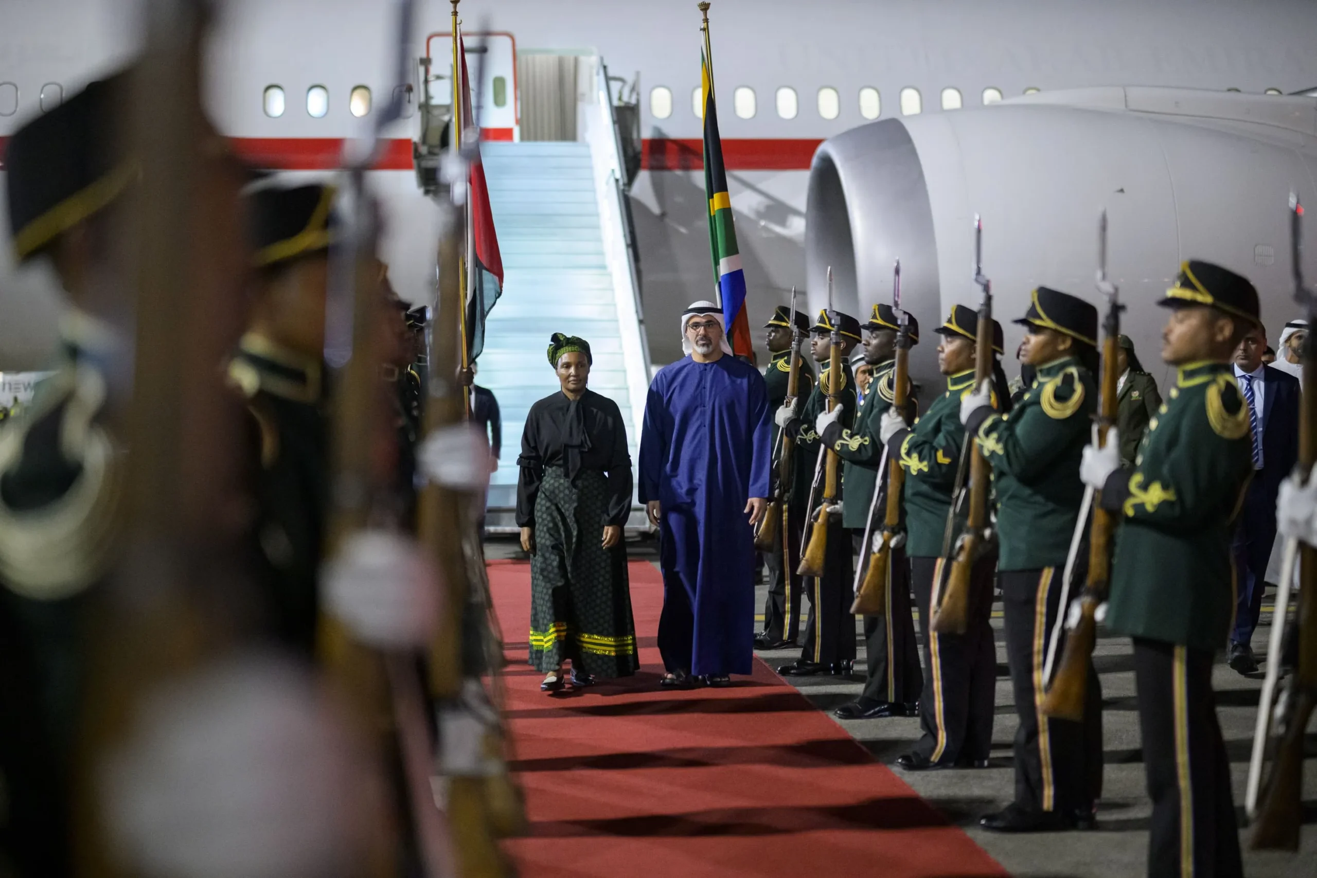 Sheikh Khaled bin Mohamed bin Zayed Al Nahyan walking on a red carpet at Johannesburg airport flanked by a military honor guard.