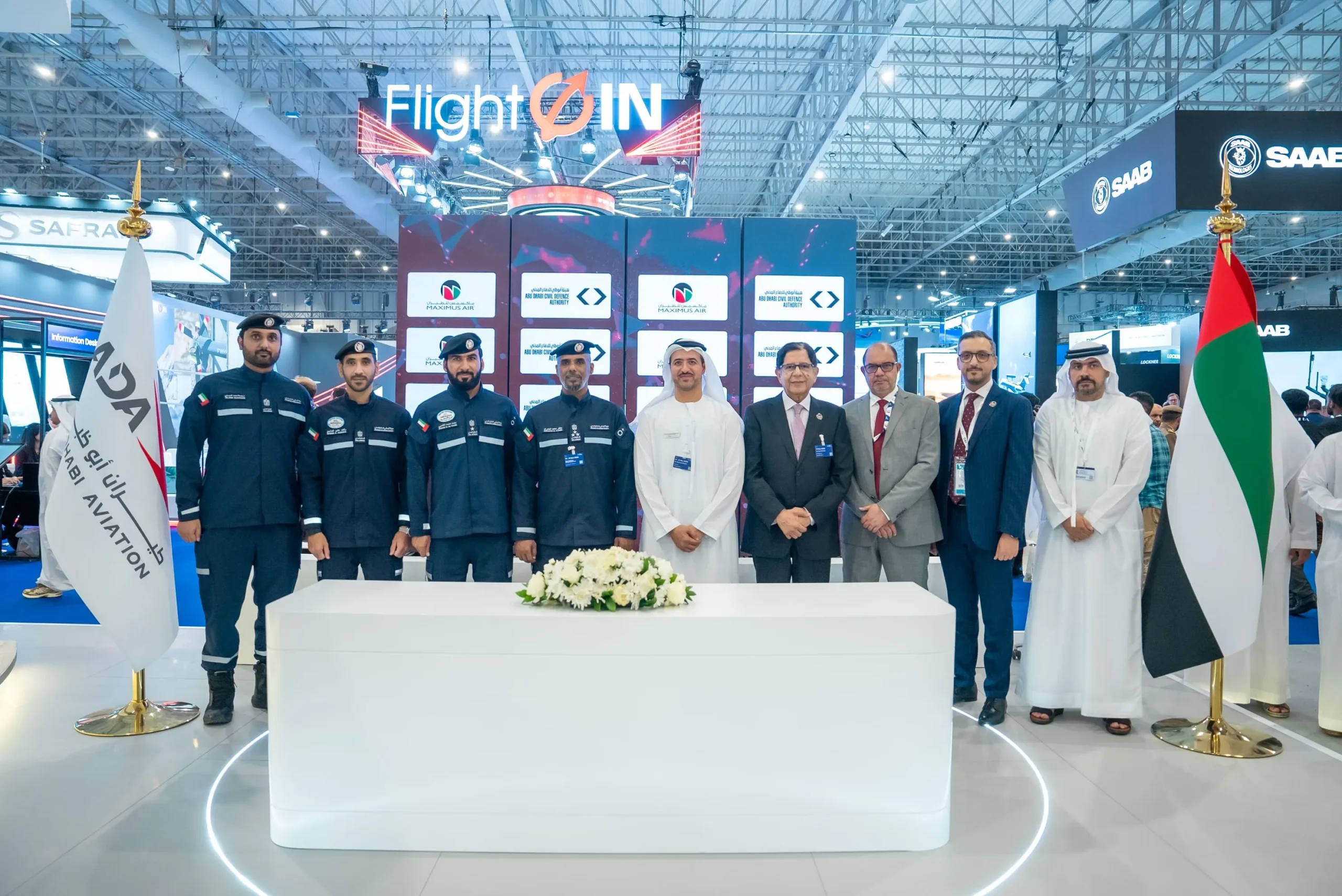 Officials from the Abu Dhabi Civil Defence Authority and Maximus Air pose for a group photo at the Dubai Airshow 2025, standing behind a white table with flowers, flanked by the Abu Dhabi Aviation and UAE flags.