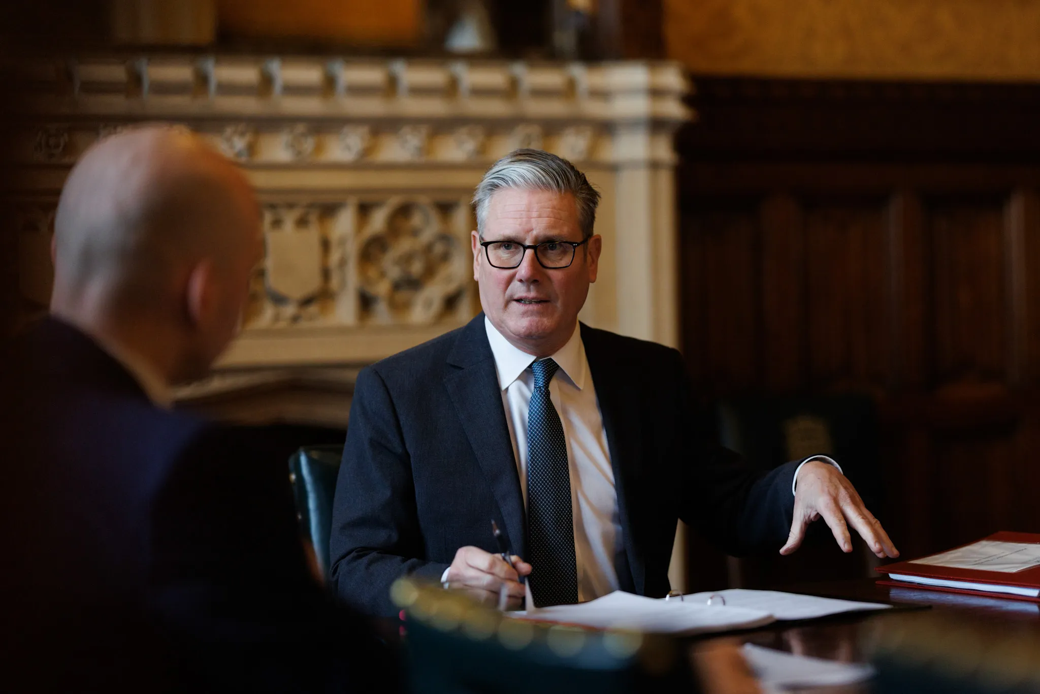 UK Prime Minister Keir Starmer sits at a polished wooden table wearing a dark suit and glasses, gesturing with his left hand while holding a pen during a formal meeting.