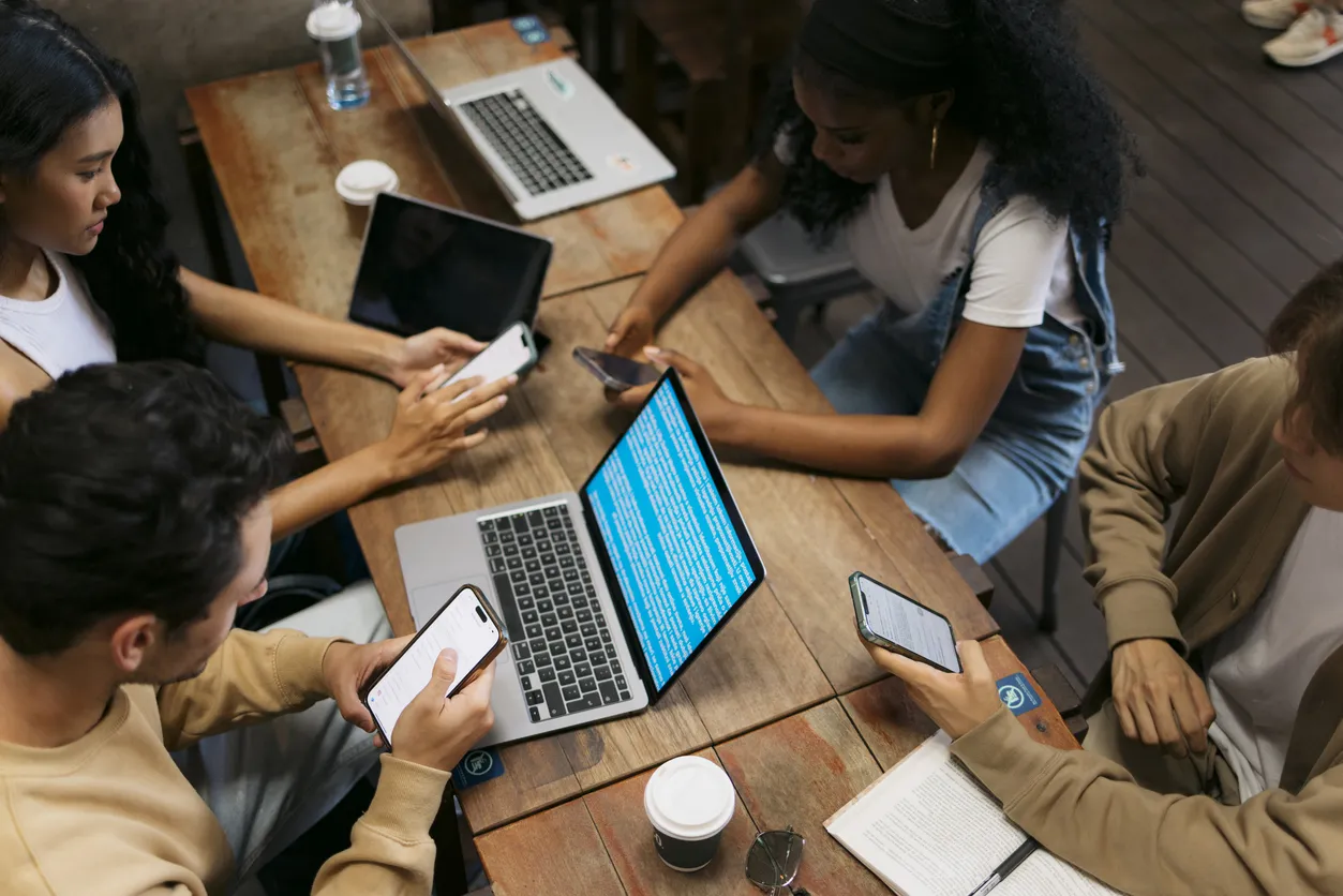 High-angle view of diverse young adults sitting at a wooden table in a cafe setting, actively using laptops and smartphones to access online platforms and digital services.