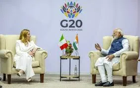 Prime Minister Narendra Modi and Italian Prime Minister Giorgia Meloni seated in armchairs holding bilateral talks on the sidelines of the G20 Summit 2025 in Johannesburg, South Africa, with national flags on a table between them.