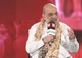 Union Home Minister Amit Shah speaking into a microphone wearing a white kurta and beige patterned shawl against a red background.
