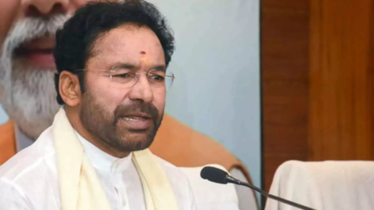 Union Minister of Coal and Mines G. Kishan Reddy addressing an event, wearing a white shirt and cream shawl, with a microphone in the foreground and a blurred poster visible in the background.