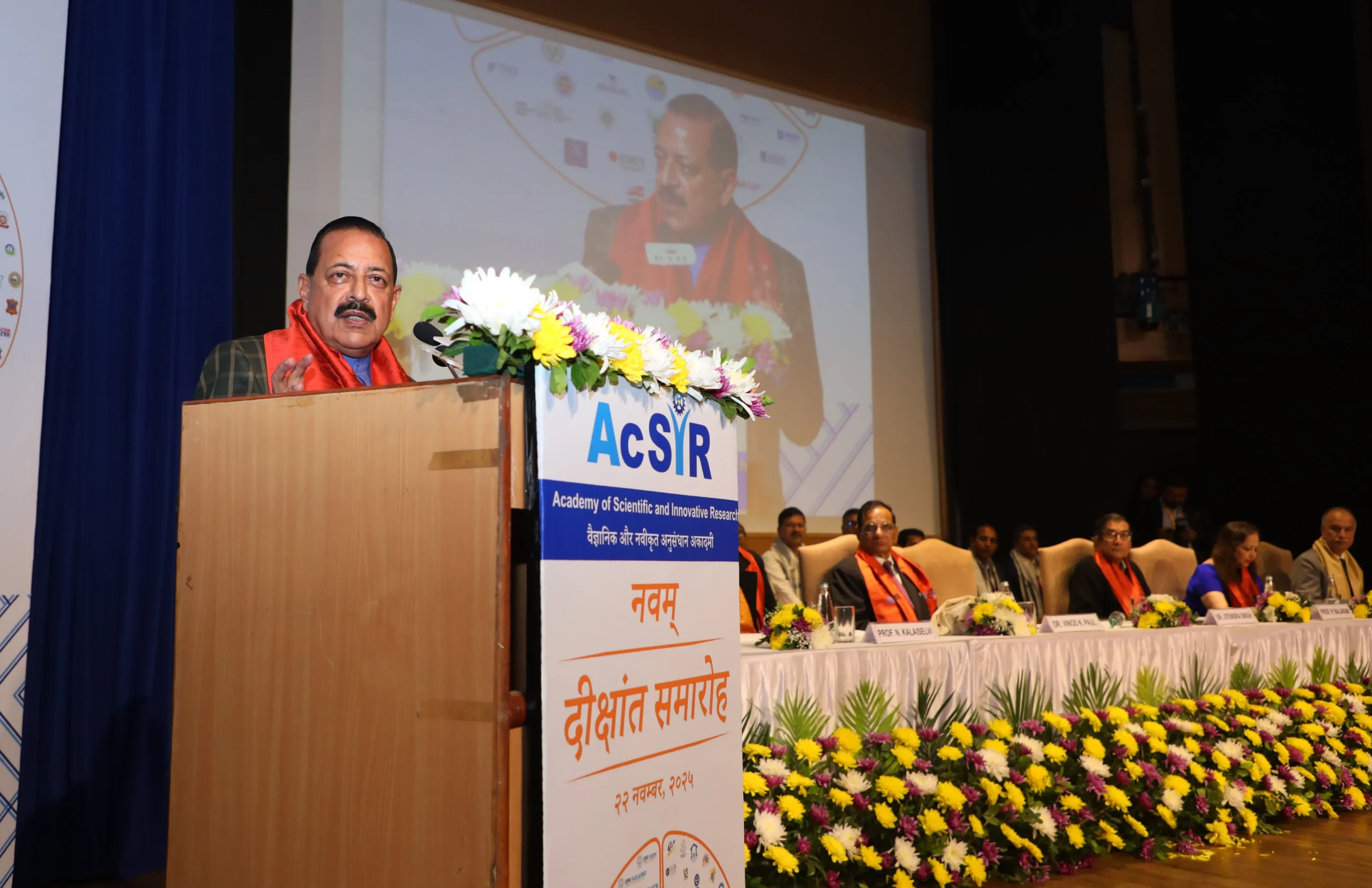 Union Minister Dr. Jitendra Singh stands at a wooden podium decorated with white and yellow flowers, delivering a speech. He is wearing a dark jacket with a red scarf. The podium features the blue "AcSIR" logo and text in Hindi and English for the "Academy of Scientific and Innovative Research" and "9th Convocation." Behind him, a large screen displays a close-up of his face, and several dignitaries wearing orange and red sashes are seated on the stage.