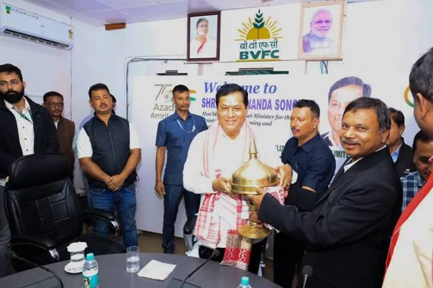 Union Minister Sarbananda Sonowal receiving a traditional brass souvenir and Gamosa during his visit to the BVFCL office in Namrup, Assam, with official portraits of PM Modi and President Murmu visible in the background.