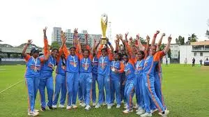 The Indian Blind Women's Cricket Team raising a gold trophy and celebrating on a green cricket field after winning the inaugural T20 World Cup.
