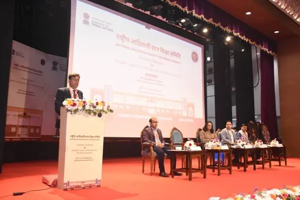 A speaker addresses the audience at the NESTS workshop on building quality infrastructure for tribal education in New Delhi, with dignitaries seated on stage.