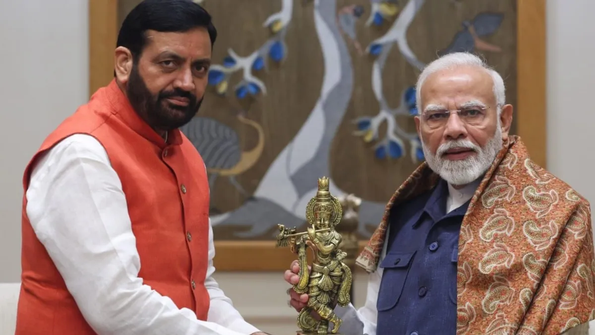 Prime Minister Narendra Modi, wearing a patterned brown shawl, and Haryana Chief Minister Nayab Singh Saini, in an orange jacket, holding a golden statue of Lord Krishna playing the flute.