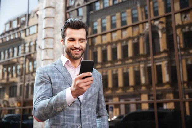A man in a grey plaid suit smiling while looking at his smartphone outdoors with a blurred city building facade in the background.