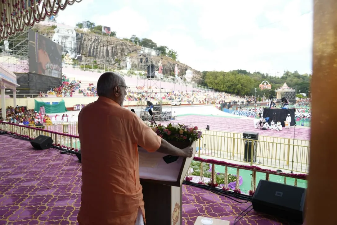 Vice President C.P. Radhakrishnan delivering a speech from a podium overlooking a large crowd at the Sri Sathya Sai Hill View Stadium in Puttaparthi during the centenary celebrations.
