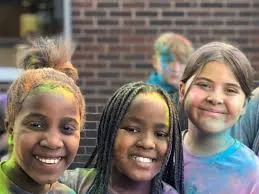 Close-up of three smiling children covered in colorful powder, featuring two girls in the foreground and a boy blurred in the background against a brick wall. 