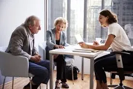A senior couple sits at an office desk consulting with a female professional who is explaining details from a document held in her hand.