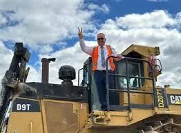 Regional Development Minister Shane Jones stands on top of a yellow Caterpillar D9T bulldozer, wearing a white shirt, tie, and orange high-visibility vest, while making a victory sign with his right hand against a cloudy blue sky.