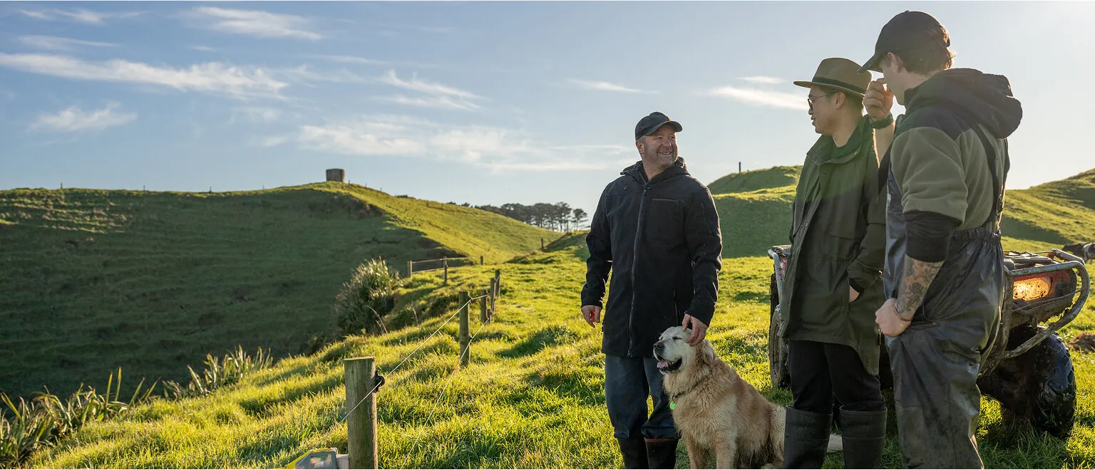 Three men standing in a green pasture on a New Zealand farm, engaging in conversation next to a quad bike; one man pets a golden dog while the group smiles, set against rolling hills and a blue sky.