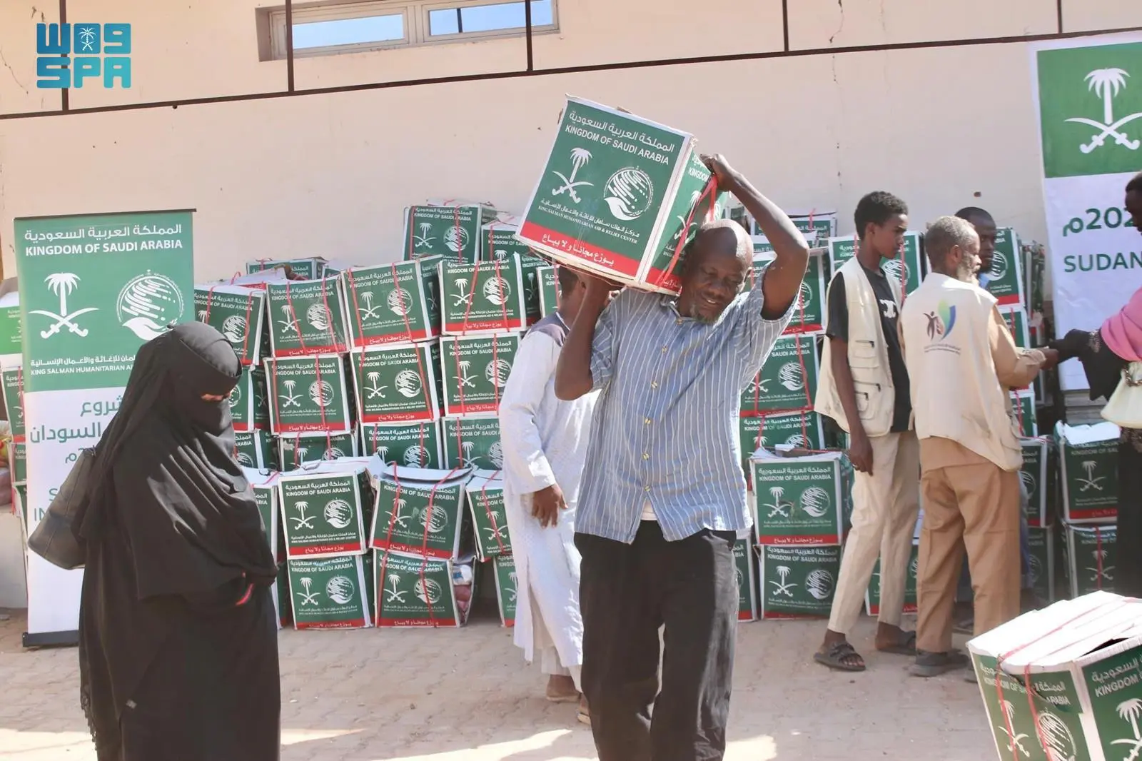 A man carries a heavy food aid box labeled 'Kingdom of Saudi Arabia' and 'KSrelief' on his shoulder, while a woman in a niqab and other beneficiaries wait near stacks of supplies in Omdurman, Sudan.