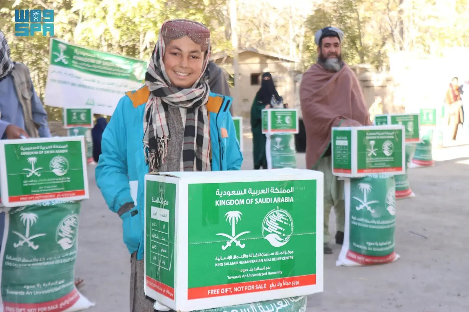 A smiling young Afghan boy in a blue jacket and traditional scarf stands beside a KSrelief food aid box during a distribution event in Baghlan Province, Afghanistan, with other beneficiaries visible in the background.