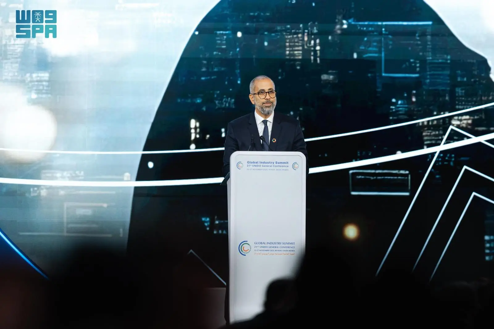 UN Resident Coordinator Mohamed El Zarkani stands at a podium delivering a speech during the 21st UNIDO General Conference in Riyadh, backed by a digital graphic display.