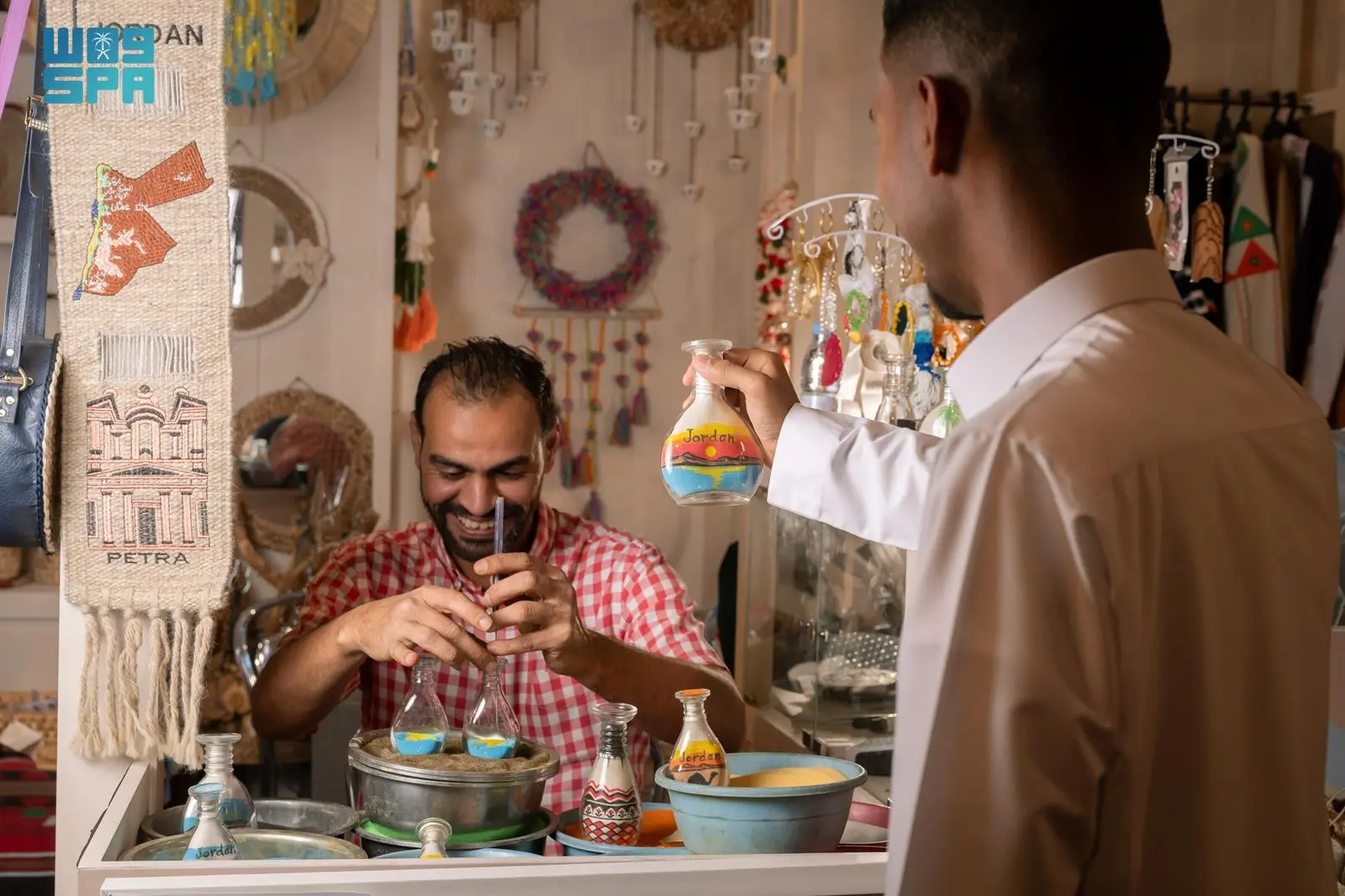 Jordanian artisan Yazan Eid filling a glass bottle with colored sand while a visitor holds up a finished souvenir featuring a desert scene at the Saudi International Handicrafts Week.