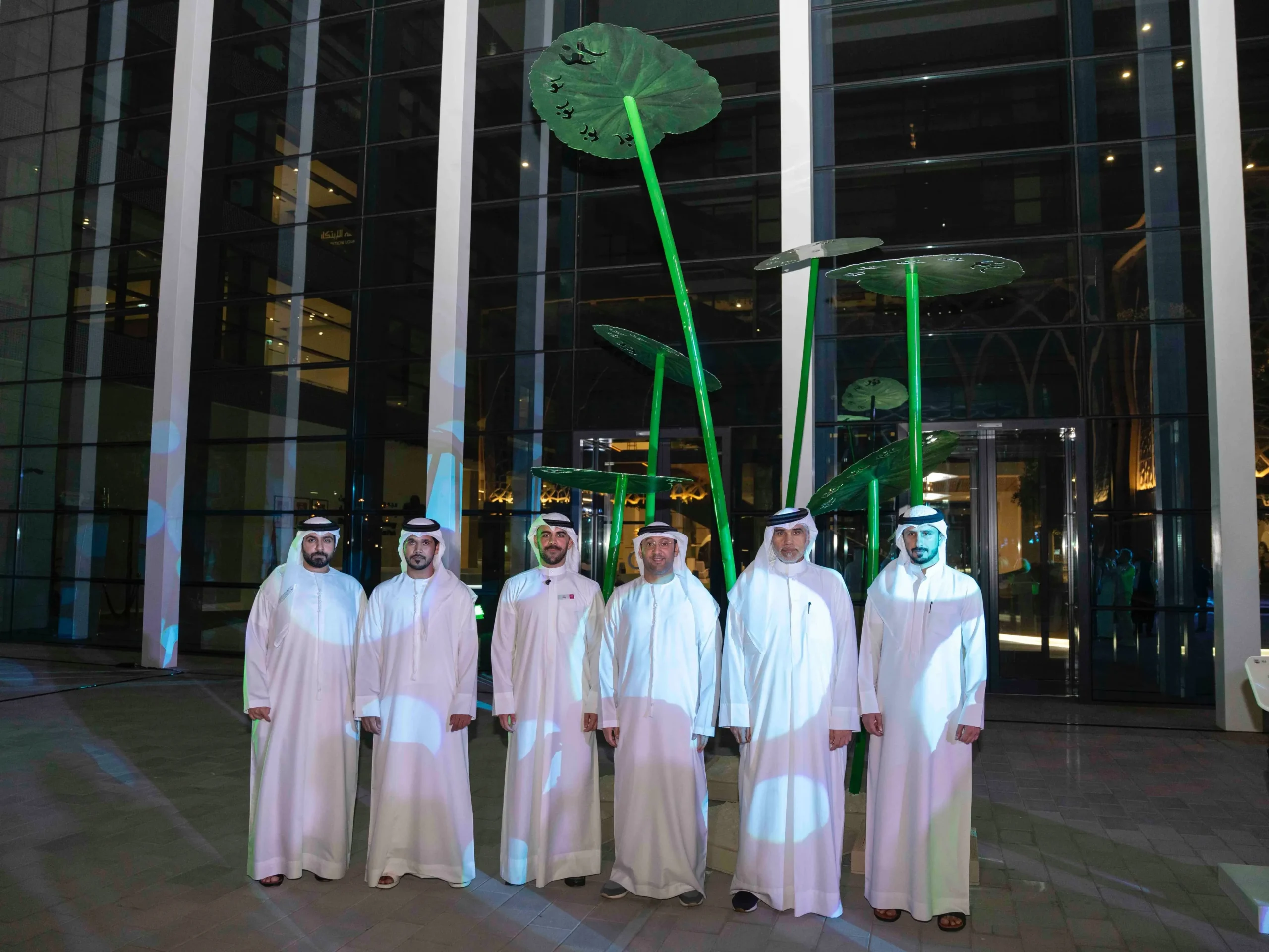 Six men in traditional Emirati white kanduras standing in front of a large green metal sculpture resembling lotus leaves with Arabic calligraphy at night.