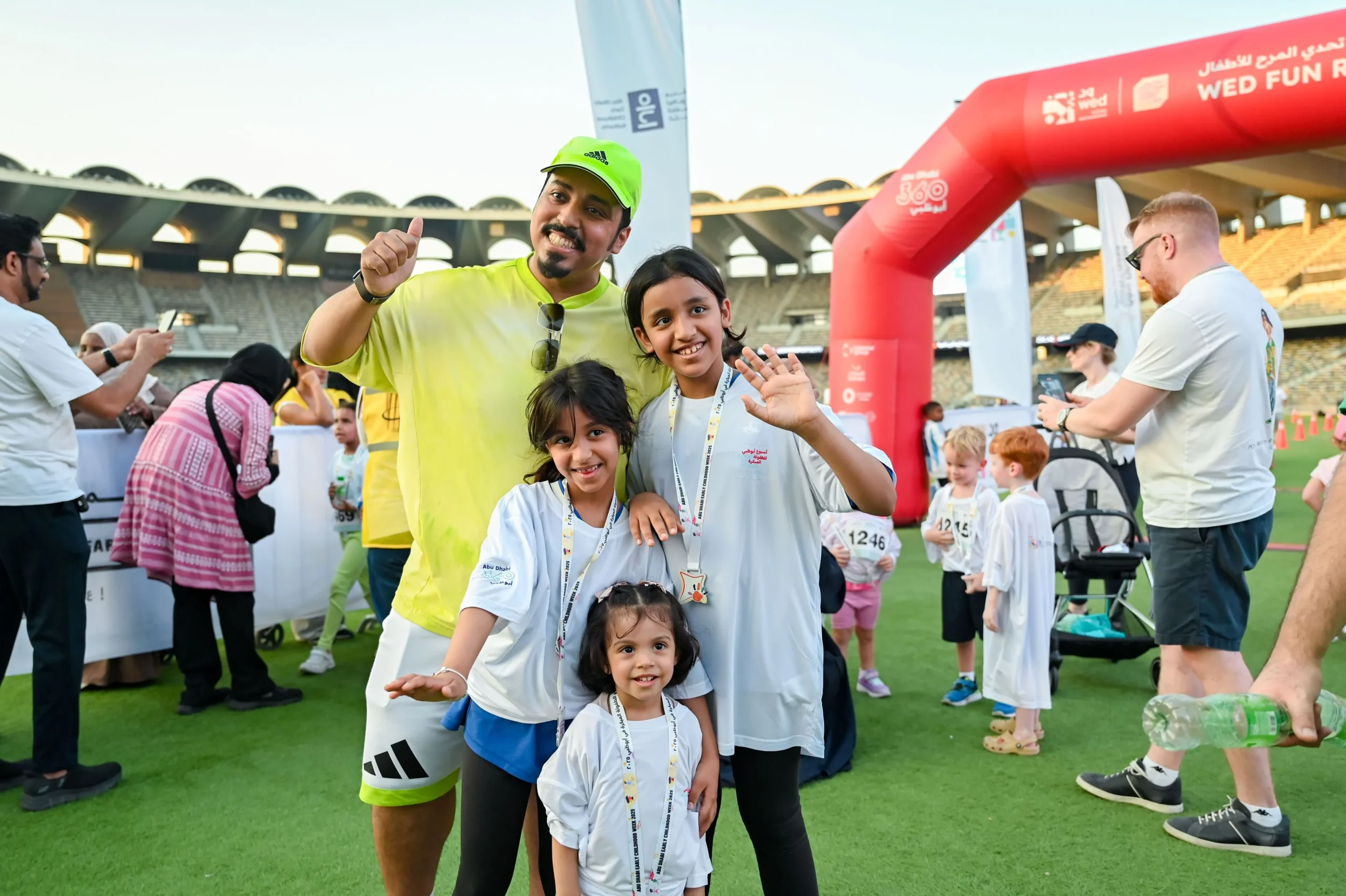 A smiling man in a neon yellow shirt and cap gives a thumbs-up while posing with three young girls wearing medals and white t-shirts at the WED Fun Run event in Zayed Sports City Stadium.
