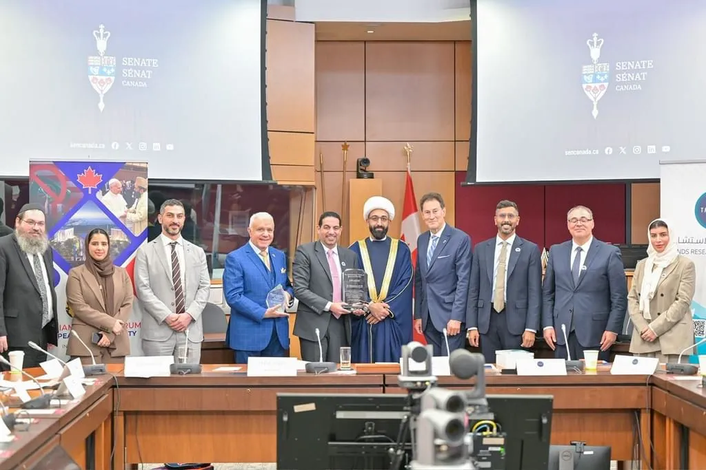 A diverse group of ten religious leaders, politicians, and researchers pose for a group photo behind a conference table in the Canadian Senate committee room. In the center, TRENDS CEO Dr. Mohammed Abdullah Al-Ali holds a glass honorary award while standing next to Imam Mohammed Tawhidi, who is wearing traditional blue robes and a white turban. Digital screens in the background display the Senate of Canada logo.