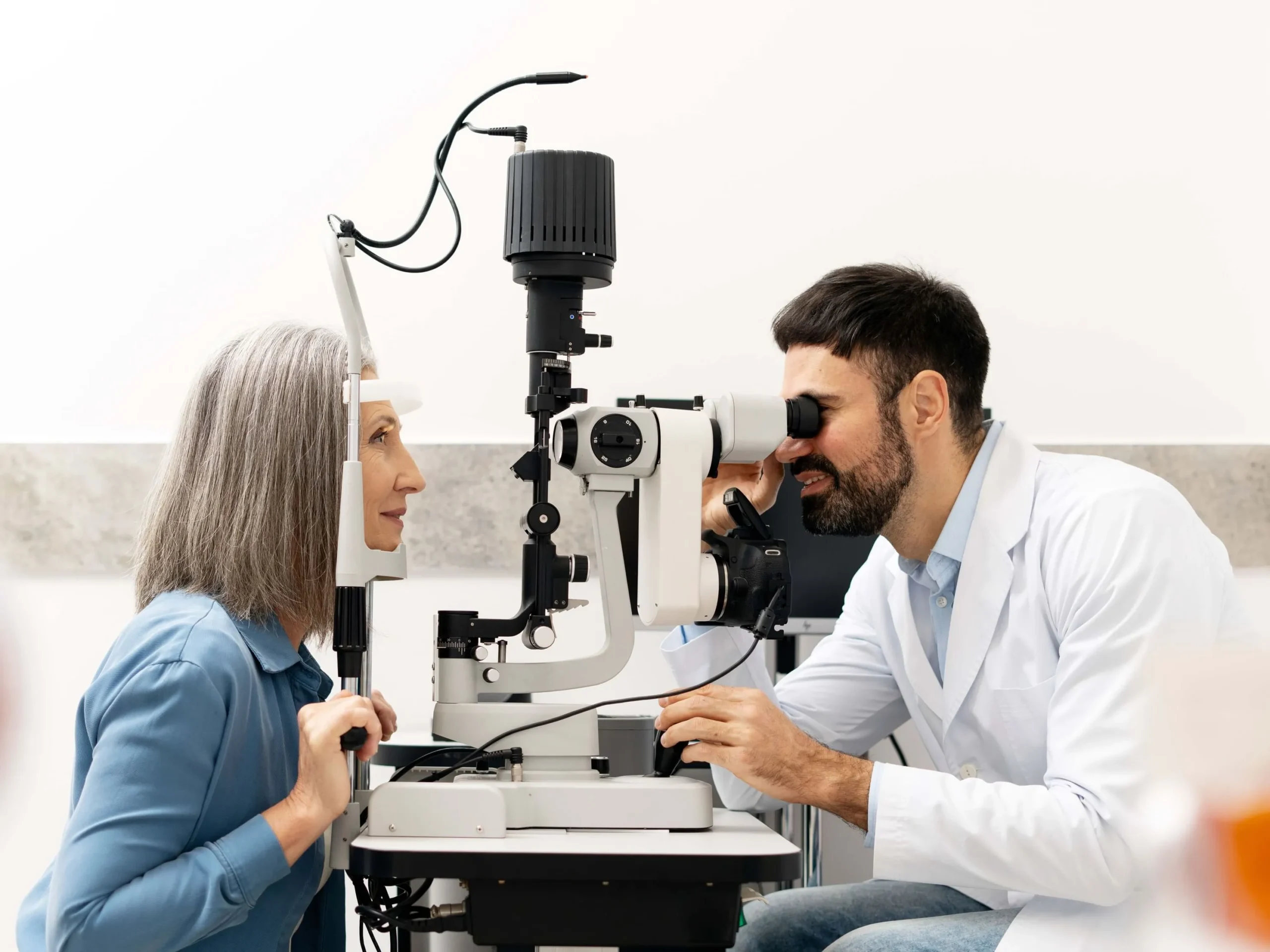 Male ophthalmologist examining a senior female patient's eyes using a slit lamp biomicroscope in a bright clinical office.