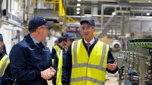 Business and Trade Secretary Peter Kyle wearing a blue cap and jacket speaks with a factory manager in a yellow high-visibility vest near a bottling line inside a manufacturing facility.