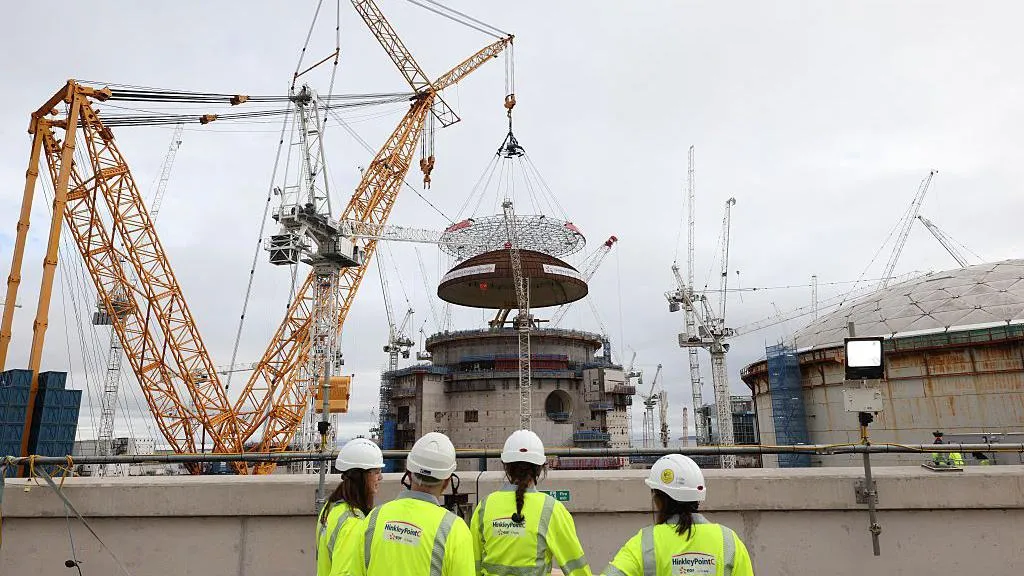 Workers wearing hard hats and Hinkley Point C high-visibility vests stand on a viewing platform watching a large yellow crane lift a massive circular steel structure onto a reactor building at a nuclear construction site. 
