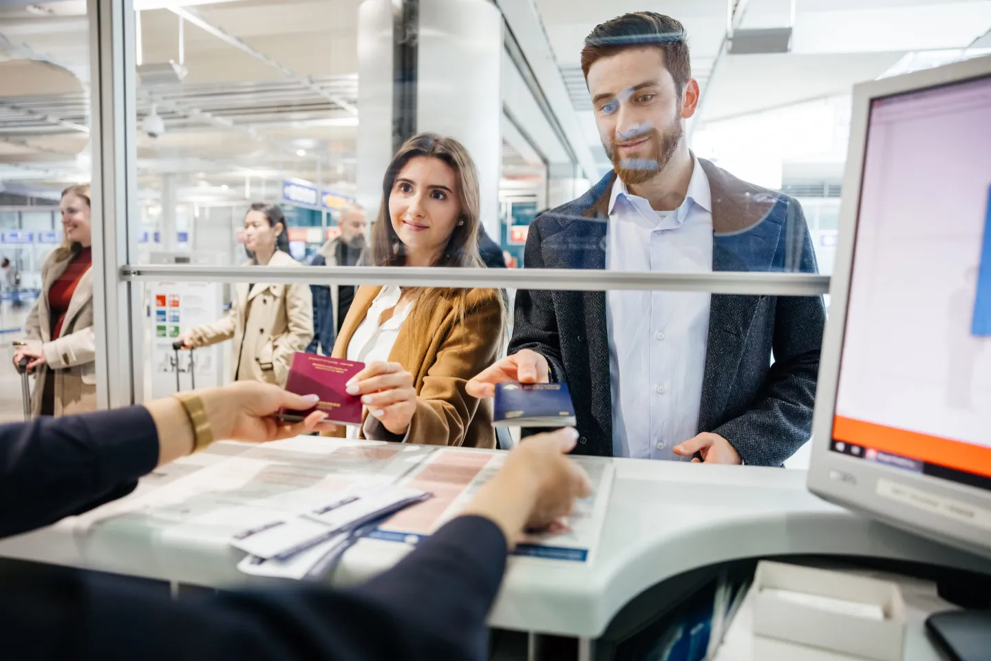 A young couple standing at an airport check-in counter handing their passports to an airline or immigration agent behind a glass partition.