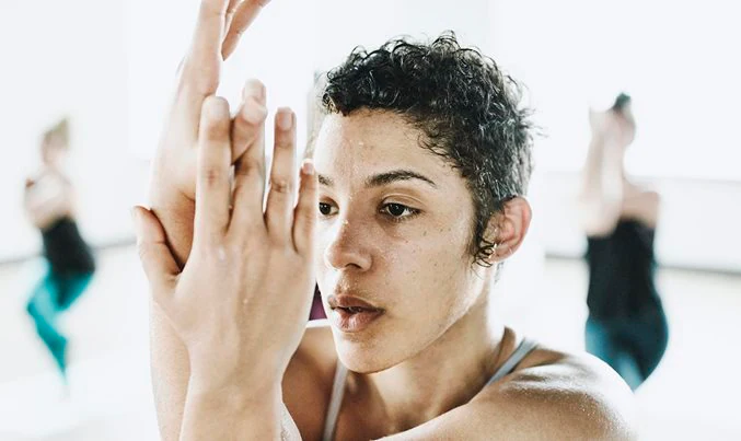 A person with short curly hair sweats while performing the Eagle Arms yoga pose during a group exercise class, illustrating focus and physical activity.