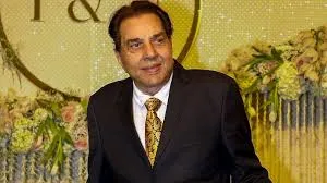 Veteran Bollywood actor Dharmendra smiling in a black suit and gold tie against a festive floral background.