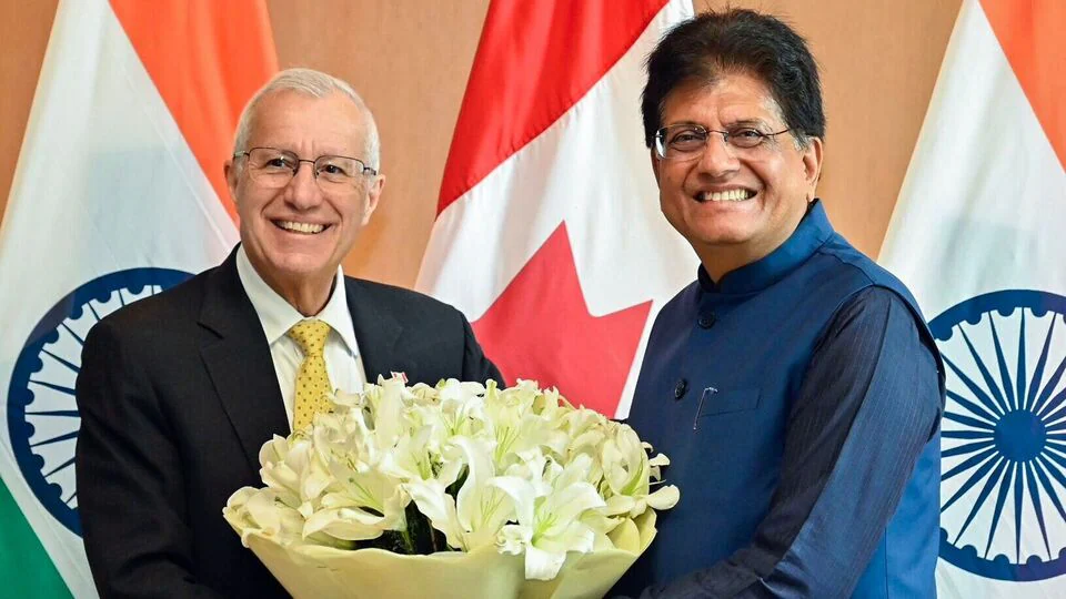 Union Minister Piyush Goyal (right) and Ontario Minister Victor Fedeli (left) smiling and holding a bouquet of white lilies during a bilateral meeting in New Delhi, with Indian and Canadian flags in the background.