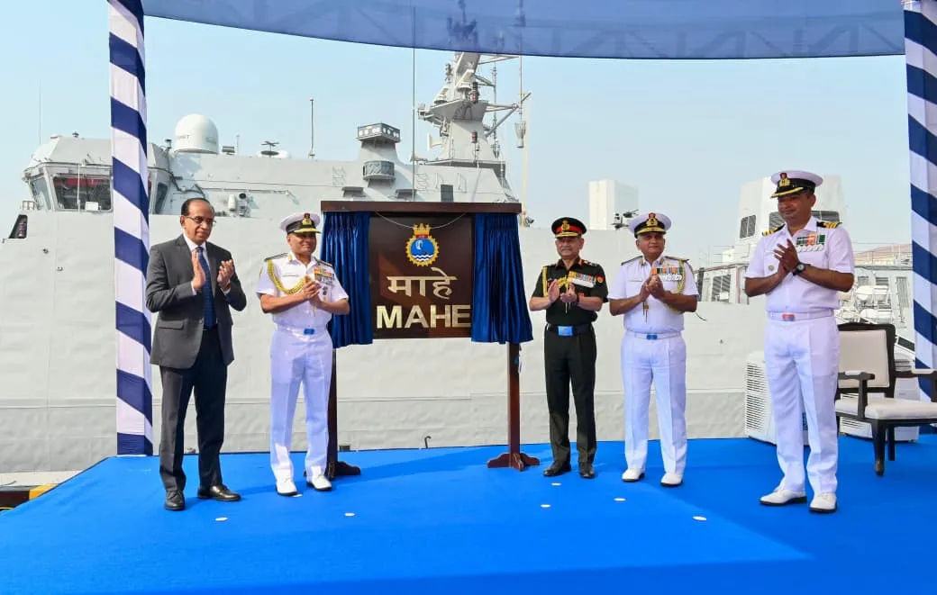 General Upendra Dwivedi and senior naval officers applaud the unveiling of the nameplate for INS Mahe during its commissioning ceremony at the Naval Dockyard, Mumbai, with the anti-submarine warfare craft visible in the background.