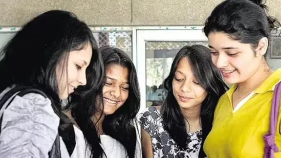 Group of four young female students smiling and looking down at a shared document or object while standing together on a school or college campus.