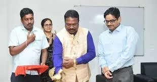 Four people standing in an indoor setting during an official event: a man in a white shirt with his right hand on his chest, a woman in a blue saree clapping her hands, a central man in a beige vest over a blue shirt with hands clasped in front, and a man in a blue shirt with hands together; a red cloth lies on the floor in front, with a whiteboard and black chair in the background.