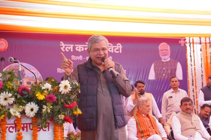Union Minister Ashwini Vaishnaw, wearing a grey vest over a kurta, speaks into a microphone while gesturing with his right hand at a podium during a railway inauguration event. Behind him is a purple banner with Hindi text reading "Rail Minister Delhi - Shamli" and a large portrait of Prime Minister Narendra Modi in a saffron kurta. The stage features floral arrangements of marigolds and white daisies, orange marigold garlands, and seated dignitaries in traditional Indian attire including saffron and white kurtas. A microphone stand and Indian Railways emblem are visible on the left.