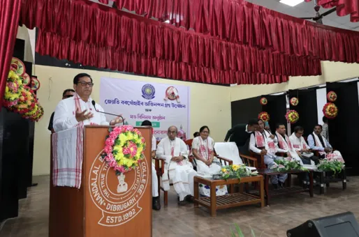 Union Minister Sarbananda Sonowal stands at a wooden podium adorned with yellow marigold flowers, delivering a speech in traditional white dhoti-kurta and gamcha. Behind him, a banner in Assamese reads the 86th Central Foundation Day of Sadou Asom Motok Sanmilan, with red curtains and floral decorations. Seated dignitaries in white attire and traditional headgear are visible on stage at Bokpara Public Auditorium in Dibrugarh, Assam.