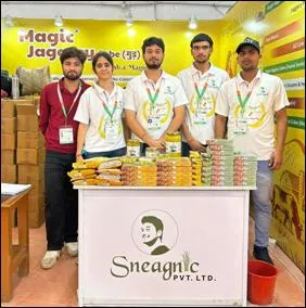 Five young adults, including three men and two women, stand smiling behind a white display table at a trade fair stall laden with stacked yellow packets of Magic Jaggery, glass jars of honey, and cardboard boxes of organic products. The yellow backdrop features the Magic Jaggery logo, while the table front displays a circular portrait of a bearded young man and the Sneha Organic Pvt Ltd nameplate. All wear white shirts with green lanyards and badges, set against a bustling exhibition hall with brown flooring and red buckets.