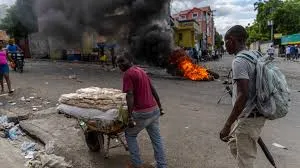 A man in a red shirt pushes a wooden cart loaded with white sacks along a debris-strewn urban street, while thick black smoke billows from orange flames of burning barricades in the background; another man in beige pants stands nearby with a backpack, and several pedestrians including a woman in pink and a motorcyclist are visible amid colorful buildings and scattered trash in what appears to be a Haitian neighborhood.