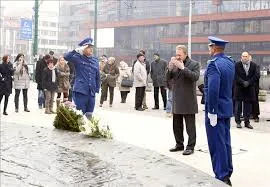 US Ambassador salutes while laying a wreath at the Eternal Flame monument in Sarajevo during Bosnia and Herzegovina Statehood Day ceremony, with Bosnian honor guards in blue uniforms present.