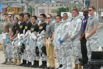 A line of multinational personnel in white protective hazmat suits and blue gloves stand on a dock at a Japanese port, with cargo ships, coastal buildings, and a yellow crane in the background under overcast skies.