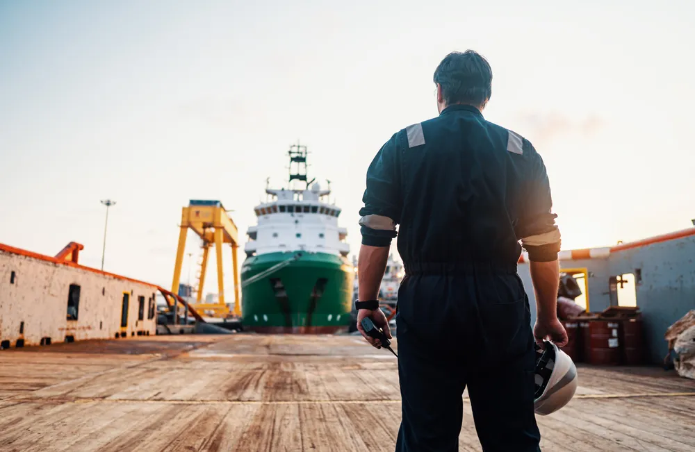Back view of a male maritime worker in black high-visibility coveralls, holding a yellow hard hat in one hand and a handheld radio in the other, standing on a weathered wooden dock at sunset, facing a large green-hulled offshore support vessel docked at an industrial port with a yellow gantry crane, shipping containers, and barrels in the foreground and background.