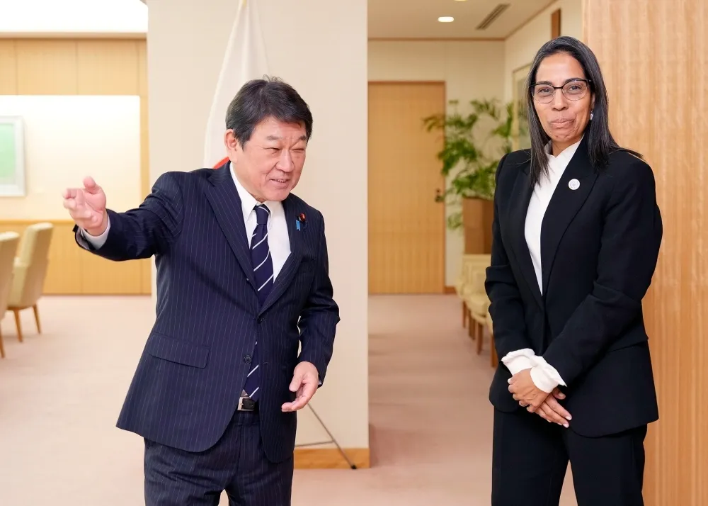 Japanese Foreign Minister Toshimitsu Motegi, in a dark pinstripe suit with a striped tie, gestures welcomingly with his right hand while smiling, stands beside Letícia Reis de Carvalho, Secretary-General of the International Seabed Authority, who wears a white blouse, black suit jacket, and glasses, with hands clasped in front, in a formal wooden-paneled office featuring a Japanese flag, potted plant, and armchairs.
